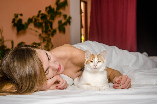 mooie jonge vrouw ligt in een wit bed met een gember kat, glimlacht, kust en knuffelt haar, liefde dieren, vriendschap - Foto, afbeelding