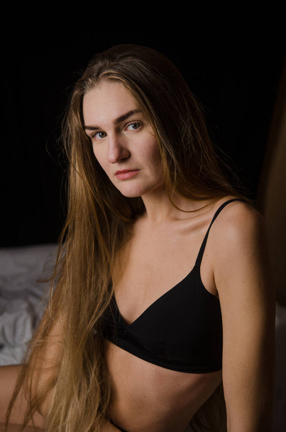 Красивая молодая женщина с светлыми волосами в черном белье сидит на кровати в эстетических позах, на черном фоне - Фото, изображение