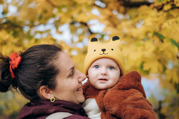 Осенью в парке гуляет женщина со своим ребенком. Молодая женщина держит своего ребенка на руках и стоит под деревом. Семейные прогулки осенью в парке. - Фото, изображение