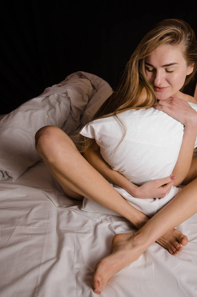 Hermosa mujer joven delgada con el pelo largo y rubio en una cama blanca se sienta abrazando una almohada y se ríe - Foto, Imagen