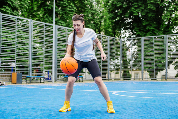 Concept of sports, hobbies and healthy lifestyle. Young athletic woman is training to play basketball on modern outdoor basketball court. - Photo, Image
