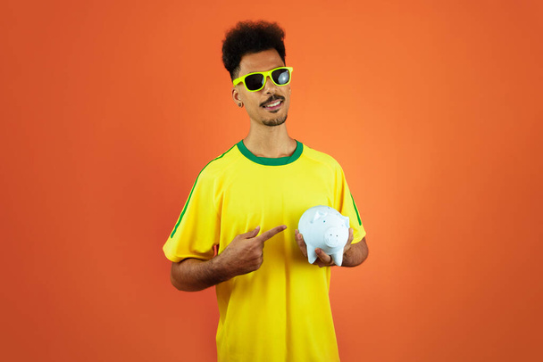 Soccer Player - Black Man Celebrating Holding Pig Coin Isolated on Orange - Photo, Image