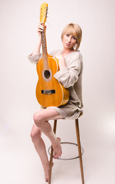Genç sarışın Bayan gri süveterli sandalyede oturan ve akustik gitar çalmak - Fotoğraf, Görsel