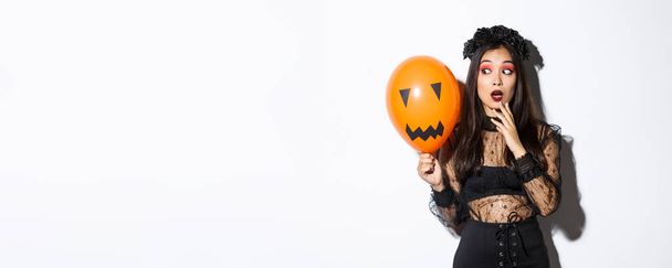 Portrait of girl looking scared at orange balloon with creepy face, wearing witch costume, celebrating halloween. - Photo, Image