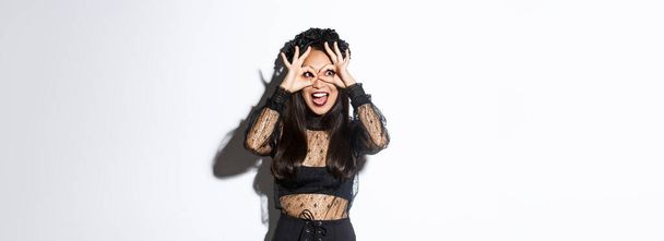 Amazed beautiful asian woman in halloween costume looking through finger glasses with excited happy face, standing over white background wearing gothic witch dress. - Photo, Image