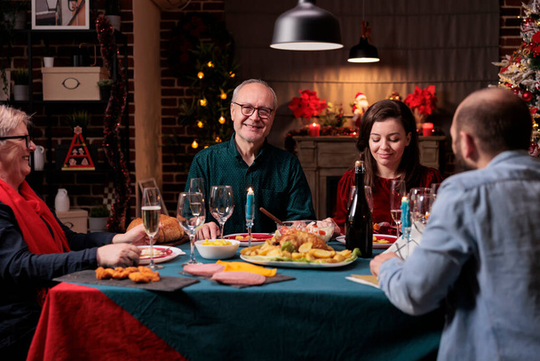 Wielka rodzina świętuje Boże Narodzenie razem, jedząc domowe potrawy przy świątecznym stole na przyjęciu domowym. Uroczystości Bożego Narodzenia w pięknym, urządzonym miejscu z ciepłym światłem świecy - Zdjęcie, obraz