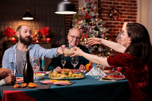 Velká rodina slaví Vánoce, jí tradiční jídlo u slavnostního jídelního stolu, muž předává ženské misky s jídlem. Vánoční zimní dovolená oslava s rodiči na krásném zdobeném místě - Fotografie, Obrázek