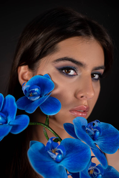 Dikey göz alıcısı, büyüleyici, baştan çıkarıcı esmer kadın mavi makyajlı ve boynundaki çiçeklerle siyah stüdyoda kameraya bakıyor. Parfüm endüstrisi, yumuşak tat. Kozmetik Parfümü - Fotoğraf, Görsel