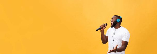 A vidám pozitív sikk portréja. jóképű afrikai férfi kezében mikrofon és fejhallgató fején hallgat zenét ének dal élvezi hétvégi nyaralás elszigetelt sárga háttér. - Fotó, kép