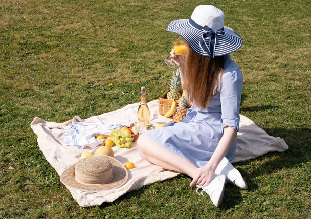 a young woman in a blue dress and straw hat is resting on a picnic with fruits, cheese plate and champagne, rest from worries and household chores parks and recreation areas,.High quality photo - Foto, Imagem