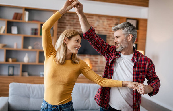 Cheerful mature european female and male relaxing, enjoy free time, dancing together, have fun in cozy living room interior. Date, love and romance, relationship, entertainment at home due covid-19 - 写真・画像