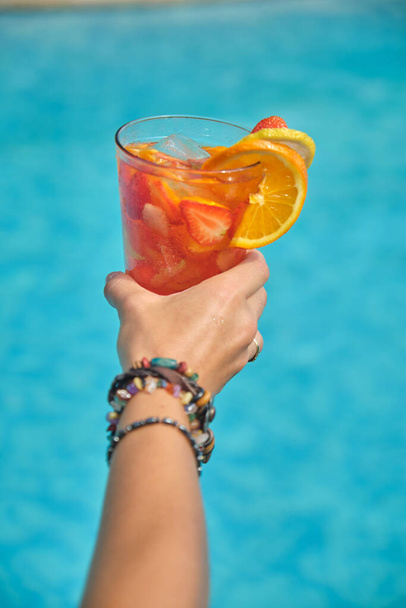 dámské ruce se sklenicí ovocného koktejlu s pomerančem a jahodami na pozadí gulubské vody v bazénu - Fotografie, Obrázek