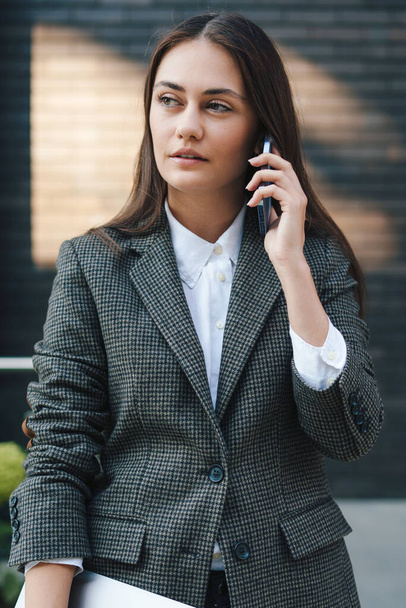 Retrato de uma mulher caucasiana atraente falando ao telefone, segurando laptop, de pé no jardim do café na cidade. Estilo de vida das pessoas. Rede de comunicação. - Foto, Imagem