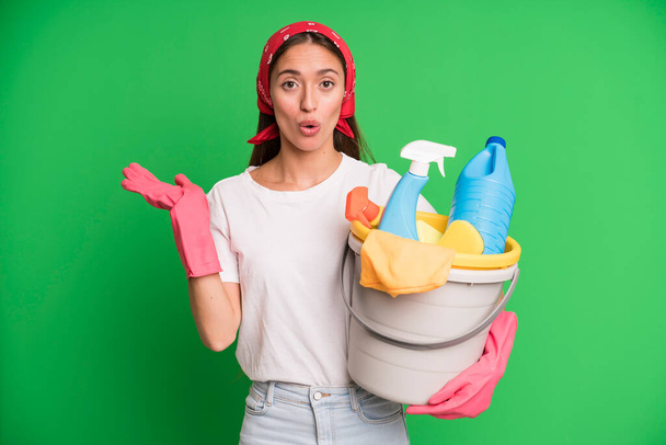 young pretty woman looking surprised and shocked, with jaw dropped holding an object. housekeeper and cleaner concept - Foto, Imagem