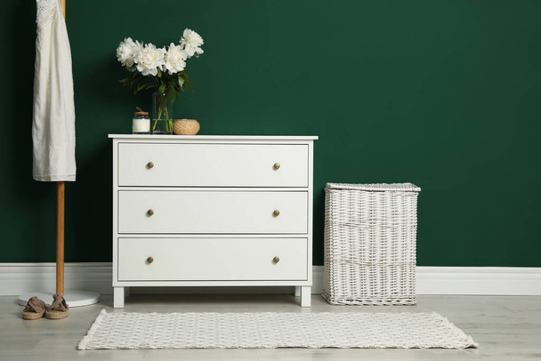 Modern white chest of drawers, coat stand and wicker basket near green wall indoors - Foto, Bild
