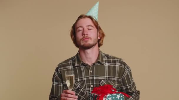 Radosny młody człowiek z tańczącym pudełkiem prezentów, świętujący szampana pijącego szklankę. Impreza urodzinowa. młody facet chłopiec zadowolony z najlepszy prezent bonus izolowane na beżowy studio tło - Materiał filmowy, wideo