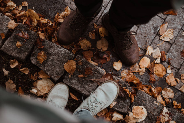 Dos pares de pies en botas de pie en la acera de pavimentación cubierta de hojas caídas. Mujer y hombre de pie juntos en el camino pavimentado con hojas de otoño alrededor. - Foto, Imagen