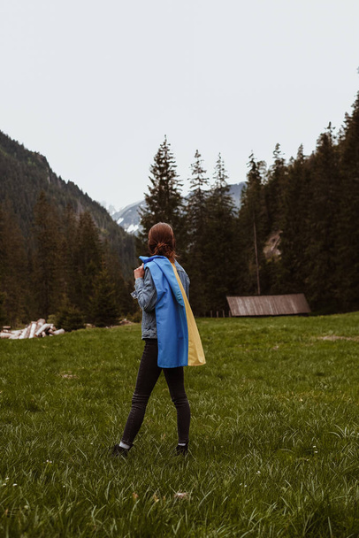 Girl in denim jacket holding Ukrainian flag on her back. Patriotic shots of a girl with Ukrainian flag in blue and yellow colors in mountains area with green grass and trees around - Photo, Image