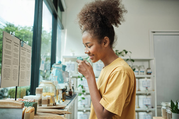 Young African American woman smells organic soaps, chooses and shops for reusable appliance products in refill store, zero-waste grocery, and plastic-free, environment-friendly, sustainable lifestyle. - Photo, Image