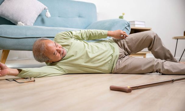 Asian old man lying on floor after falling down with wooden walking stick, Sick senior man beside couch on rug in living room at home, Elderly having an accident after doing physical therapy alone - Фото, изображение