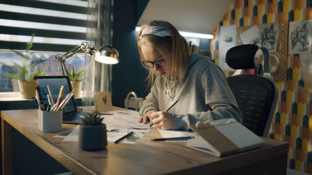 Ev yapımı bir tasarım stüdyosunda çalışan sarışın bir kadın. Kapüşonlu kapüşonlu kadın kalem çizimleri yapıyor. Video için yol haritası olarak çerçeve oluşturuluyor. - Fotoğraf, Görsel