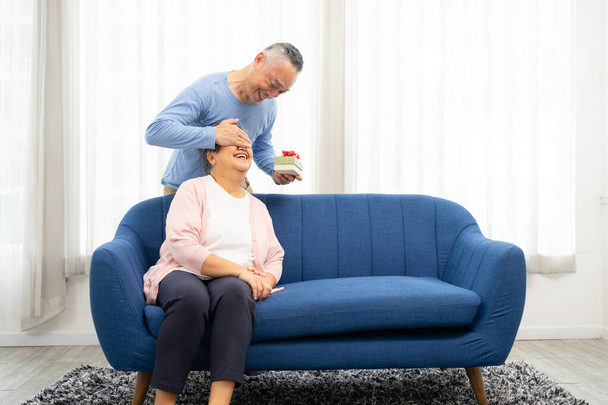 Happy Senior Asian man covering eyes of senior Asian woman for surprise with gift box in home.  Elderly Asian couple on valentine's day concept. - Photo, Image