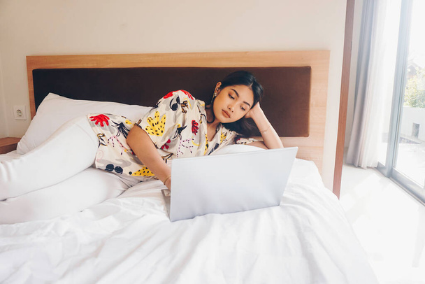 Серьезная сконцентрированная молодая женщина, работающая с постели. Держите ноутбук на коленях и набирайте на клавиатуре. - Фото, изображение