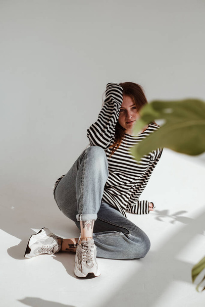 Studio portrait of red hair girl sitting on white background in black and white striped sweater and blue jeans with green plant nearby. Studio portraits of red hair girl with variety of emotions and styles. - Фото, зображення