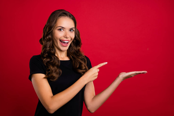 Photo of cute excited young woman wear black t-shirt holding arm pointing empty space smiling isolated red color background. - Фото, зображення