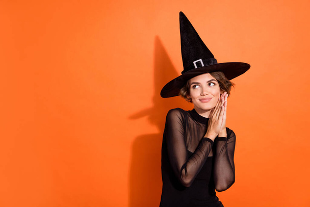 Photo of thoughtful cute lady wear black witch dress cap looking empty space isolated orange color background. - 写真・画像