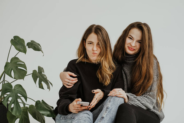 Birbirlerini kucaklayan, gülümseyen ve surat yapan iki genç kadının stüdyo fotoğrafı. Yan yana oturan iki kadının portreleri. Yanında bir canavar bitkisi, sarılıyor, duygularını ve duygularını ifade ediyor.. - Fotoğraf, Görsel