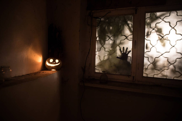 Τρομακτική κολοκύθα αποκριών στο μυστικιστικό παράθυρο του σπιτιού τη νύχτα ή κολοκύθα αποκριών και σιλουέτα τρόμου στο παράθυρο. Επιλεκτική εστίαση - Φωτογραφία, εικόνα