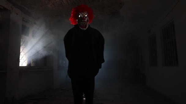 Pauroso clown malvagio con i capelli rossi in piedi all'interno sala rovinata in edificio abbandonato di notte. Focus selettivo - Filmati, video