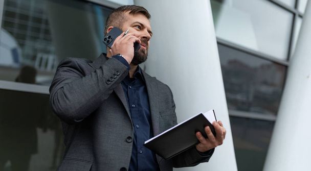 een volwassen man in zakelijke kleding spreekt op een mobiele telefoon met een notitieblok in zijn handen. - Foto, afbeelding