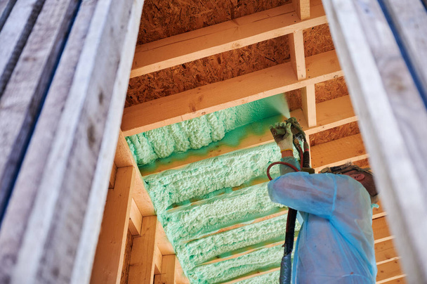 Male builder insulating wooden frame house. Man worker spraying polyurethane foam inside of future cottage, using plural component gun. Construction and insulation concept. - Foto, afbeelding