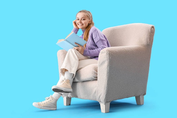 Νεαρή γυναίκα διαβάζει βιβλίο στην πολυθρόνα σε μπλε φόντο - Φωτογραφία, εικόνα