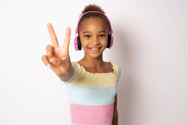 Cheerful young mixed race woman girl showing peace symbol posing isolated on white background studio portrait. People lifestyle concept. Listen music with headphones, dancing - Photo, Image