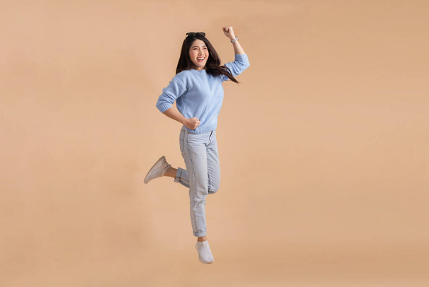 Happy smiling Asian woman with jumping in the air lifting her hands up in blue sweater and jeans isolated on beige color background. Full body length in studio shot. - Foto, imagen