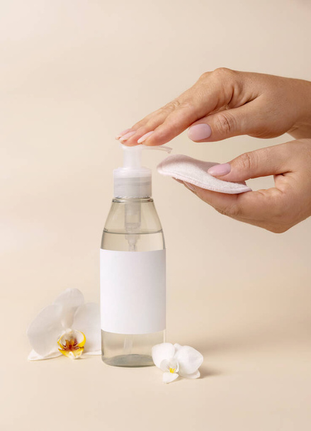 A woman hand pumping liquid on a make-up remover pad from a cosmetic bottle near orchid flowers on beige close up, label mockup. Skincare beauty product package, minimal compositio - Photo, Image