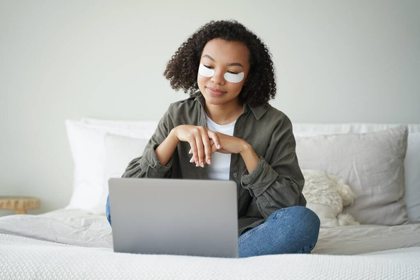 Happy afro girl applies eye patches and relaxing at home in weekend morning. Young hispanic woman having rest watching tv series. Skin moisturizing. Lovely curly girl is grooming herself in bedroom. - Photo, Image