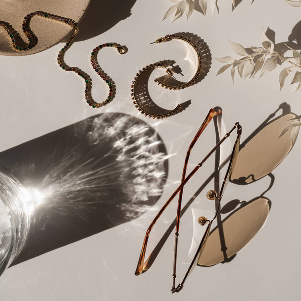 Esztétikai luxus kompozíció női ékszerekkel és kiegészítőkkel: napszemüveg, fülbevaló, karkötő fehér háttéren üveggel a napfény árnyékában. Elegáns bohém lapos fektetés, top view divat beállítás - Fotó, kép