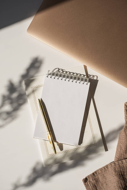 Spiraalvormige flip notebook met lege kopieerruimte, pen, laptop computer in bloemen zonlicht schaduw op witte tafel. Esthetische Boheemse minimalistische werkruimte. Artiest, schrijver sjabloon - Foto, afbeelding
