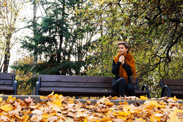 Una donna dai capelli corti si avvolge in una sciarpa seduta su una panchina in un parco autunnale - Foto, immagini