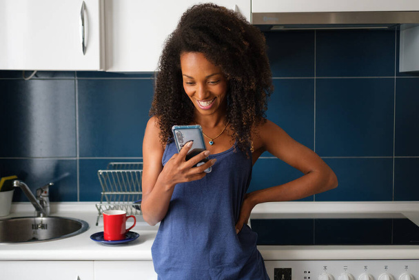 Casual afro hairstyle woman at home using smartphone for video call from the kitchen. Cheerful female searching cocking recipes online concept. - Zdjęcie, obraz