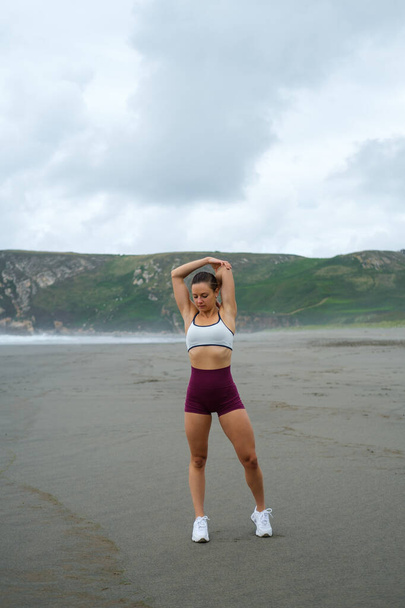 Plajda spor yapan formda bir kadın. Fitness ve sağlıklı açık hava yaşam tarzı. Kadın sporcu omuzlarını esnetiyor. Asturias kıyıları, İspanya. - Fotoğraf, Görsel