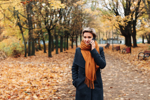 Krótkowłosa blondynka komunikuje się przez telefon spacerując w jesiennym parku w płaszczu i szaliku. - Zdjęcie, obraz
