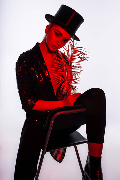 Full length image of a young woman with makeup wear in a glitter costume and top hat, sitting on chair, isolated white background. - Foto, imagen