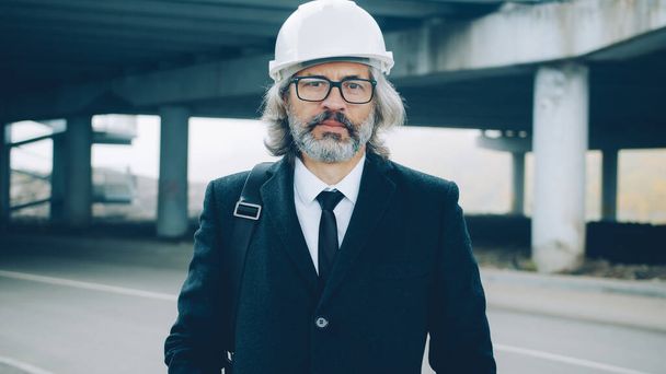 Portrait of respectful businessman standing in industrial zone outdoors wearing safety helmet and stylish suit and looking at camera with serious face - Fotografie, Obrázek