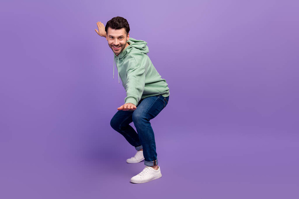 Pleine longueur photo de jeune homme hipster porter belle tenue kaki sweat à capuche denim positif calme souriant posant en vous regardant isolé sur fond de couleur violet vif. - Photo, image