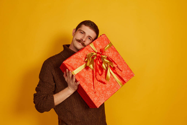 Portrait of cute young man holding big present box isolated over yellow studio background. Looks delightful and happy. Concept of holidays, happiness, emotions, facial expression, joy, celebration - Фото, зображення